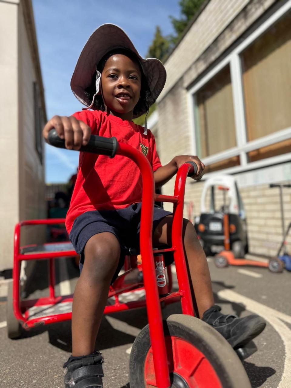 A young child in a sunhat rides a trike across the nursery playground.